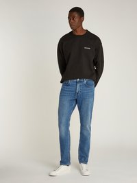 Sudadera negra con logotipo en el pecho, combinada con jeans rectos de azul medio y zapatillas blancas. El modelo está de pie con las manos detrás de la espalda.