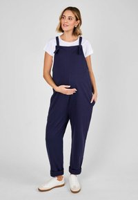Marineblaue Umstandsoveralls mit verstellbaren Trägern und Schleifenakzenten, im lockeren Schnitt, mit seitlichen Taschen und umgeschlagenen Manschetten. Kombiniert mit einem weißen T-Shirt.