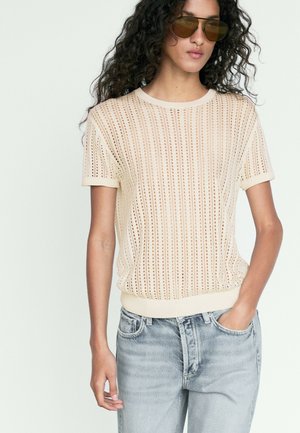 Vrouw met bruine zonnebril en een creme kort mouw gebreide top met verticale opengewerkte patronen, gecombineerd met lichtblauwe jeans.