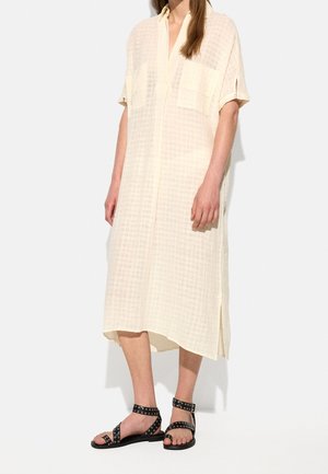 Femme portant une robe midi beige clair texturée avec des manches courtes et des sandales noires à bride cheville avec des œillets argentés.