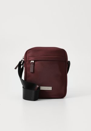 Sac bandoulière bordeaux en tissu doux avec quincaillerie en ton argent, sangle noire réglable et poche zippée à l'avant. Présente une plaque de logo.