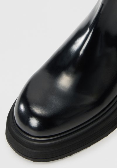 Botte en cuir verni noir avec un bout arrondi, semelle en caoutchouc noir épaisse et surface lisse et propre. Pas de motifs ni de matériel visibles.
