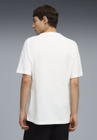 Νέος άνδρας με κοντά καστανά μαλλιά που φοράει απλό λευκό κοντομάνικο μπλουζάκι, γυρισμένος με την πλάτη, μπροστά σε γκρίζο φόντο με διαβάθμιση.