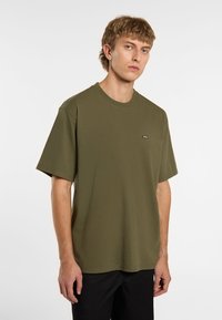 T-shirt in cotone verde oliva con collo a giro, maniche corte e una piccola etichetta logo sul lato sinistro del petto. Tessuto liscio e vestibilità rilassata.