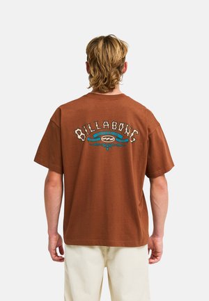 Uomo con capelli biondi che indossa una t-shirt marrone Billabong con un logo blu e beige sulla schiena, abbinata a pantaloni beige chiaro.