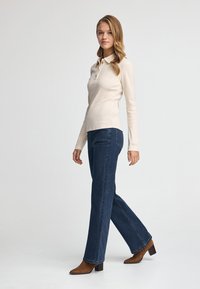 Polo beige a maniche lunghe con colletto, abbinato a jeans a zampa d'elefante blu scuro e stivaletti con tacco marroni. Design semplice, texture liscia.