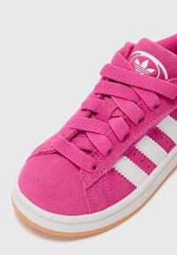 Pink suede sneaker with white leather stripes, featuring a rounded toe, textured laces, and rubber sole. Branding on the tongue.