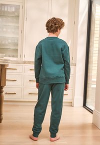 Sweatshirt bleu sarcelle et pantalon de jogging assorti en tissu doux, avec une coupe décontractée et des poignets côtelés, vus de dos. Pieds nus.