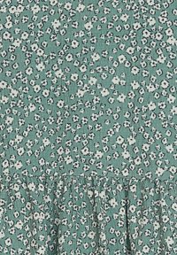 Grüner Stoff mit einem kleinen Blumenmuster in Weiß und Schwarz. Das Material hat eine strukturierte Oberfläche und verfügt am Saum über eine geraffte Rüsche.