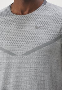 Šedé sportovní tričko Nike s texturovaným vzorem a tmavšími akcenty, s kulatým výstřihem a diskrétním logem v horní levé části hrudi.