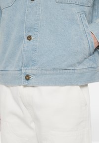 Ljusblå denimjacka med bronsknappar, som har två sidfickor, en klassisk krage och rak fåll. I kombination med vita byxor.