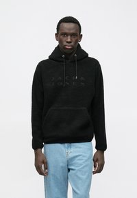 Νεαρός άνδρας φορώντας καφέ υφασμάτινη ζακέτα με λογότυπο "Jack & Jones" και ανοιχτό μπλε τζιν, σ standing μπροστά σε λευκό φόντο.