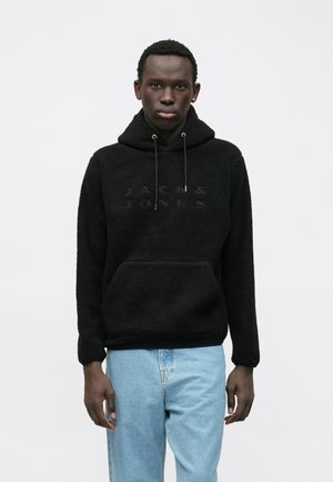 Mlad moški v črni teksturirani kapuci z logotipom "Jack & Jones" in svetlo modrimi jeans hlačami, ki stoji proti enobarvni beli ozadju.