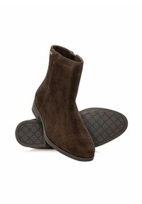 Brązowe zamszowe botki za kostkę z zaokrąglonym noskiem, niskim piętem na klocku i teksturowaną gumową podeszwą z wzorem w diamenty. Szczegół z boku zamka błyskawicznego.