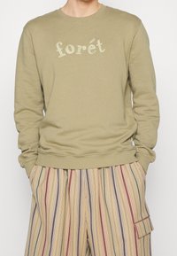 Oliwkowy sweatshirt z haftowanym napisem "forét", w luźnym kroju, z prążkowanym dekoltem i mankietami. W zestawie z beżowymi spodniami w paski.