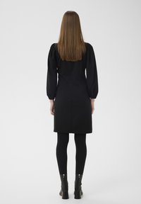 Rochie neagră cu mâneci bufante, lungime până la genunchi, croială ajustată. Poartă colanți negri și botine cu detaliu de fermoar.