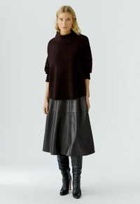 Vrouw staat gekleed in een zwart oversized sweater, zwarte leren midi-jurk en zwarte kniehoge haklaarzen tegen een effen achtergrond.