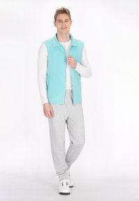 Hellblaues Zip-Up-Westen über einem weißen Langarmshirt, kombiniert mit grauen Jogginghosen und weißen Turnschuhen. Glatter Stoff mit lässiger Passform.