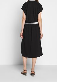 Woman wearing a black midi dress with short sleeves, a striped waistband, and black strapped heels with clear block heels, standing facing away.