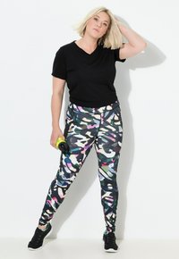 Zwarte v-hals t-shirt gecombineerd met multicolore camouflage legging met roze, blauwe en groene patronen; houdt een gele fitnessaccessoire vast.