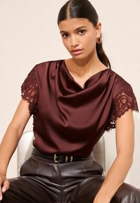 Top din satin bordeaux cu mâneci din dantelă, având un decolteu relaxat. Asortat cu pantaloni din piele maro, cu talie înaltă, și o curea neagră.