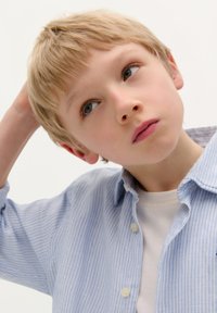Jongen met kort, lichtblond haar in een lichtblauwe en witte gestreepte overhemden over een wit t-shirt, die omhoog kijkt.