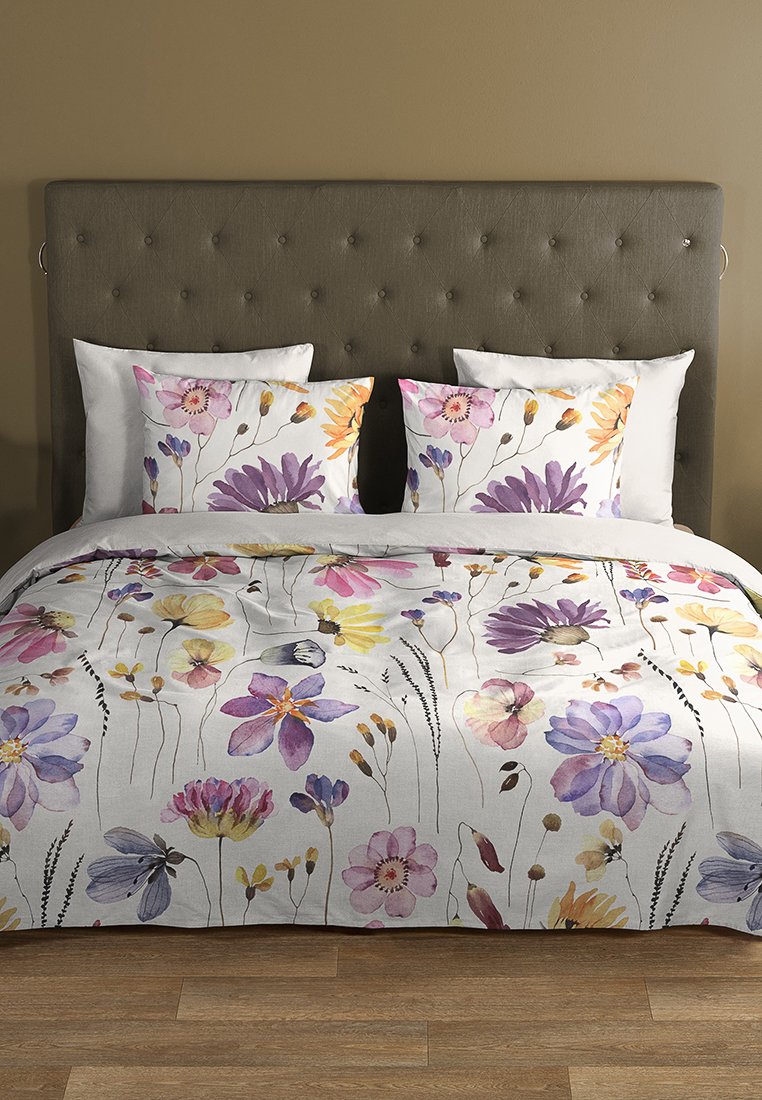 Brettbett mit dunkelgrauem gepolstertem Kopfteil, weißen und blumigen Kissenbezügen sowie passendem, blumigen Bettbezug auf Holzboden.