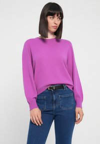 Lilla, myk strikkegenser med lange ermer og rund hals, kombinert med mørkeblå, tettsittende jeans og et svart belte med gullspenne.