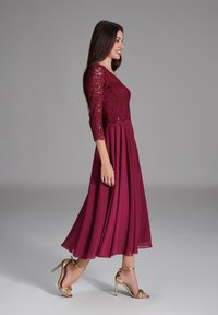 Robe en dentelle bourgogne à manches trois-quarts et jupe fluide. Associée à des chaussures à talons dorés, mettant en valeur une silhouette élancée.