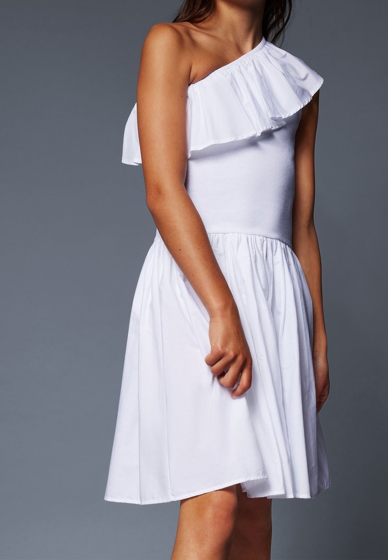 Femme en robe blanche sans manches avec un col volanté asymétrique et une jupe froncée, debout devant un fond gris.