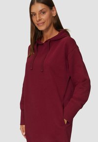 Bordeauxfarbenes Hoodie-Kleid mit einer Kängurutasche, langen Ärmeln und einer Kordelzugkapuze. Hergestellt aus weichem, einfarbigem Stoff mit lockerer Passform.