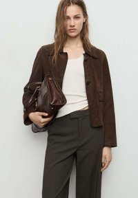 Giacca in suede marrone con colletto, chiusura a bottoni e tasca laterale. Abbinata a una maglietta bianca a maniche corte e pantaloni scuri, tiene in mano una borsa marrone.