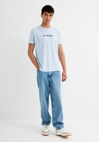 Camiseta de algodón azul claro con texto negro "TODO LO QUE VEO ES AZUL", combinada con jeans holgados azul claro y zapatillas blancas con acentos marrones.