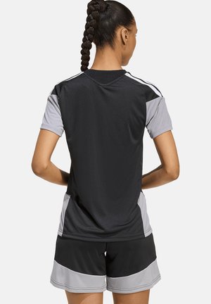 Kvinde med fletninger, iført sort og grå sportstrøje og shorts, står med hænderne på hoften, vendt væk fra seeren.