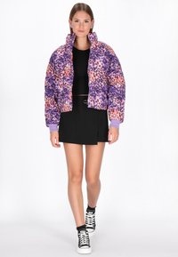 Pufová bunda s leopardím potiskem v purpurové, růžové a oranžové barvě. Má vysoký límec a žebrované manžety. Nosit s černou sukní a teniskami.