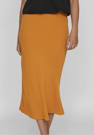 Vrouw met een mosterdgele midirok met een lichte uitloping, gecombineerd met een zwarte mouwloze top, staand tegen een effen achtergrond.