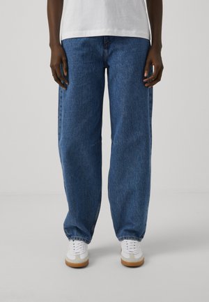 Osoba nosząca luźne niebieskie dżinsy, białą koszulkę i białe trampki stojąca na tle jednolitego jasnego tła.