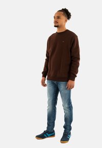 Bruine sweater, blauwe jeans en sneakers met blauwe accenten. Heeft een ronde halslijn en ribgebreide manchetten. De stof lijkt zacht en gestructureerd.