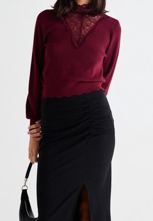 Femme portant un haut bordeaux à manches longues avec un détail en dentelle et une jupe noire texturée avec une fente à l'avant, tenant un sac à main noir.
