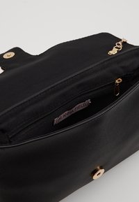 Svart handväska med en slät läderutsida, har en dragkedjeförsedd innerficka, guldfärgad beslag och en märkt etikett inuti.