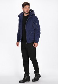 Tengerészkék párnázott dzseki, kapucnival, elülső cipzárral, két nagy foltvarrásos zsebbel és egy cipzáras ujjzsebbel, fekete ing és nadrág felett viselve.