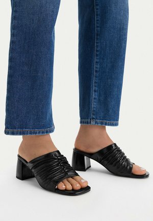 Persona che indossa sandali neri con tacco, punta aperta e arricciati, e jeans blu, in piedi su una superficie bianca.