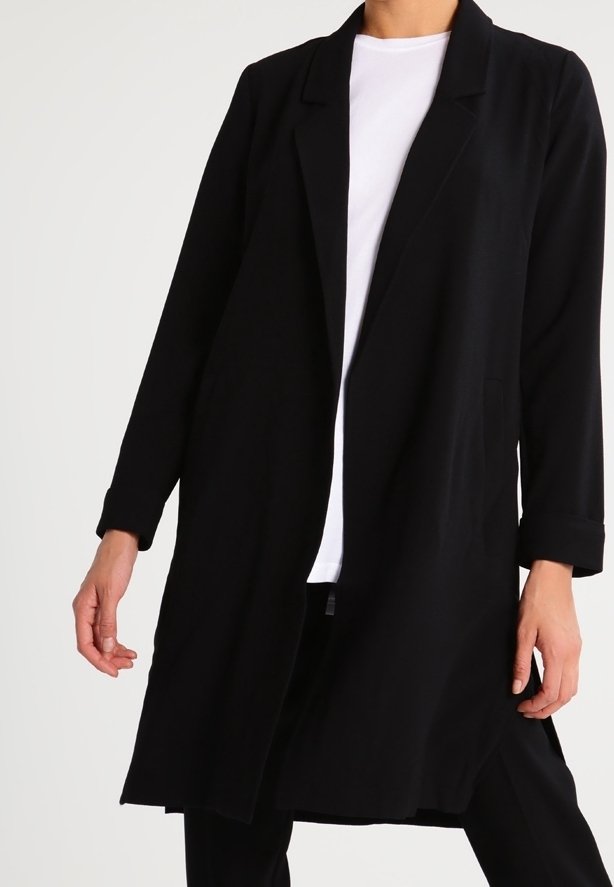 Personne portant un long manteau noir ouvert sur une chemise blanche, debout, les bras relâchés le long du corps devant un fond uni.