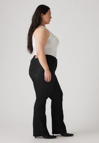 Black flared jeans with a high waist, featuring a classic five-pocket design and a leather back patch. Model wears a white tank top.