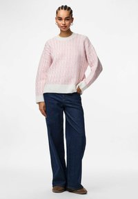 Stickad tröja med ett rosa och vitt mönster samt ribbstickade muddar och fåll. Matchad med mörkblå vidbyxade jeans och beige skor.