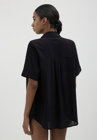 Maglietta nera a maniche corte in tessuto leggero con vestibilità ampia, colletto piatto e cucitura centrale sul retro. Maniche arrotolate e orlo leggermente curvato.