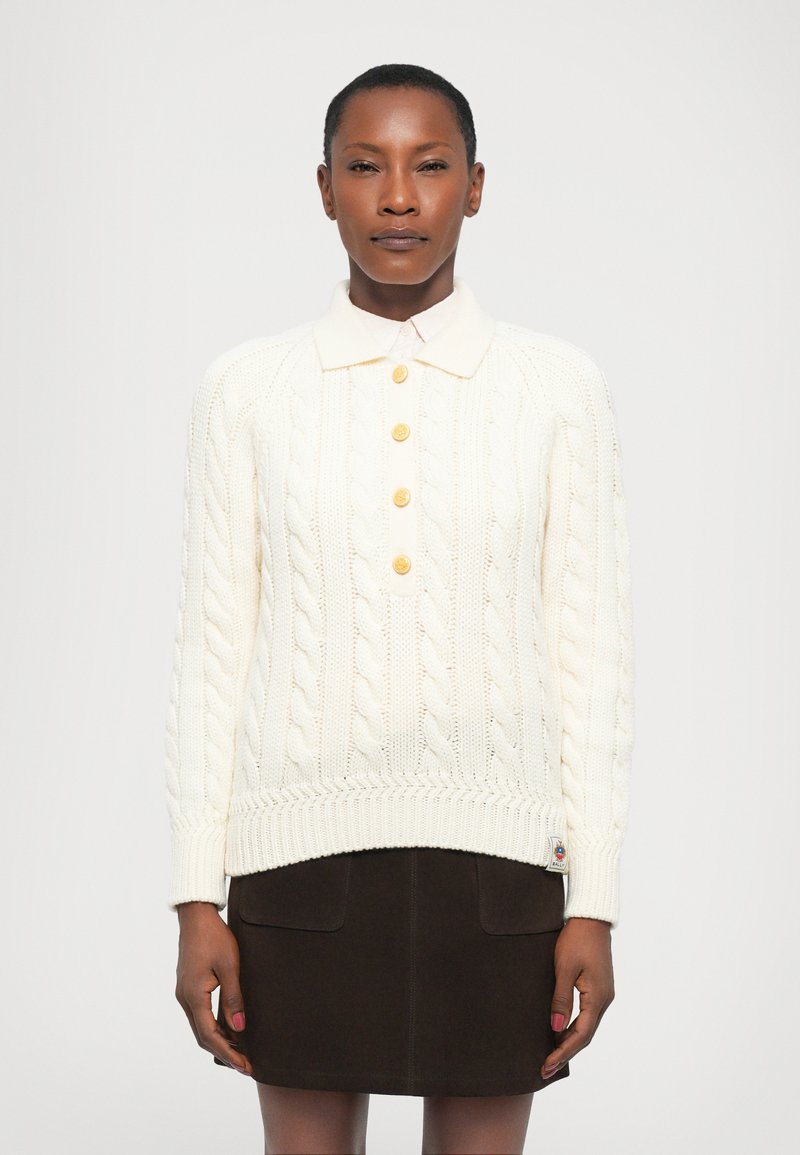 Pull en tricot torsadé crème avec un col, orné de quatre boutons dorés et de manches côtelées, assorti à une jupe marron foncé.