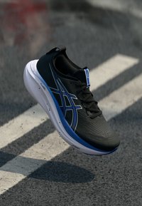 Chaussure de sport noire avec un dessus texturé, des accents bleus et une semelle intermédiaire blanche. Présente un logo de marque proéminent et une fermeture à lacets.