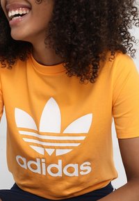 T-shirt en coton jaune avec un grand logo trefoil Adidas blanc et trois bandes horizontales. Manches courtes et col rond.