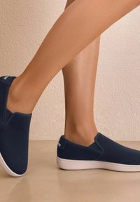 Granatowe buty slip-on z teksturowaną siateczką i białą gumową podeszwą. Posiadają elastyczne akcenty po bokach oraz logo na pięcie.